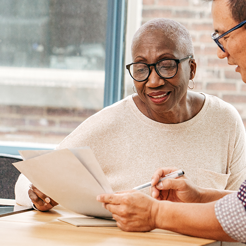An older Black woman in a weathered cream top and black glasses discusses the contents of a paper with an Asian man in a checkered shirt and black glasses. They are sitting at a light coloured wood board room table in front of a window.