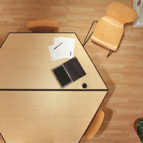 An aerial view of a simple hexagonal wood table. Chairs are placed on each side, and one is pulled out in front of a section of the table that holds a laptop, cup of coffee, assorted papers and a pen.