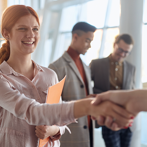 A smiling and approachable-looking young woman with red hair is holding an orange document folder in her left hand while shaking the hand of someone in front of her, who is out of view. Two men in suits are talking and smiling in the background.