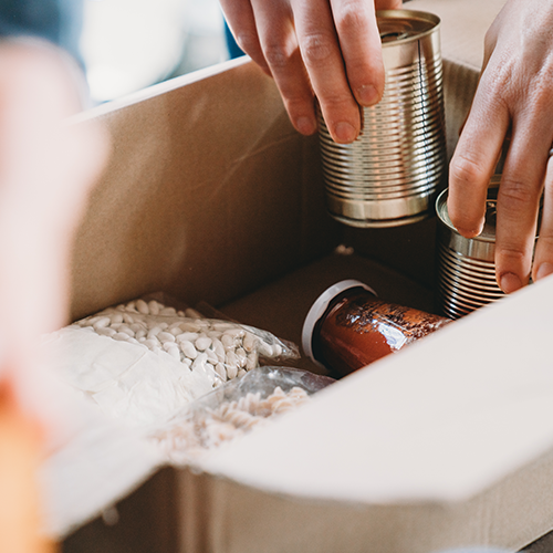 A cardboard box with a jar of sauce, package of rotini pasta, and package of dried beans. A hand is reaching inside with two unlabelled aluminum cans of food.