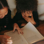 young multi-racial female children laying in bed reading a book together giggling