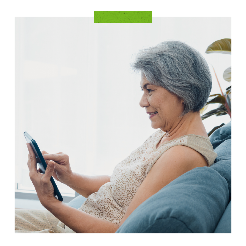 Happy Asian senior woman sit on couch, holding and looking at digital tablet screen in hand in living room.
