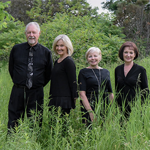 A group of four senior adults dressed in all black, standing in a row in tall grass and smiling at the camera.